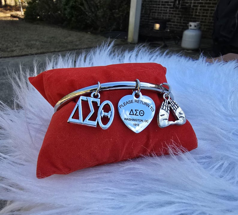 May include: Silver charm bracelet with a heart charm that says "Please return to Delta Sigma Theta Washington, DC 1913" and other charms including a Greek letter charm and a charm with two hands clasped in prayer.