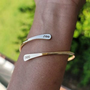 Könnte beinhalten: Ein goldfarbenes Armreif-Armband mit den eingravierten Texten "ZΦB" und "1920". Das Armband ist gebogen und offen, zum Tragen am Handgelenk konzipiert. Der Hintergrund ist eine verschwommene Ansicht von grünem Laub.