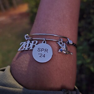 Puede incluir: Un brazalete de plata con tres dijes. Los dijes son un círculo de plata con el texto "SPR '24", un dije de plata con las letras "ZΦB" y un dije de plata en forma de pájaro con un ojo azul.