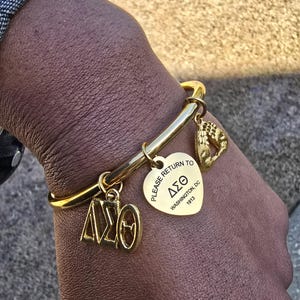 May include: Gold charm bracelet with a heart-shaped charm that says "Please return to Delta Sigma Theta, Washington, DC 1913". The bracelet also has a charm with the letters "DST" and a charm with a handprint.