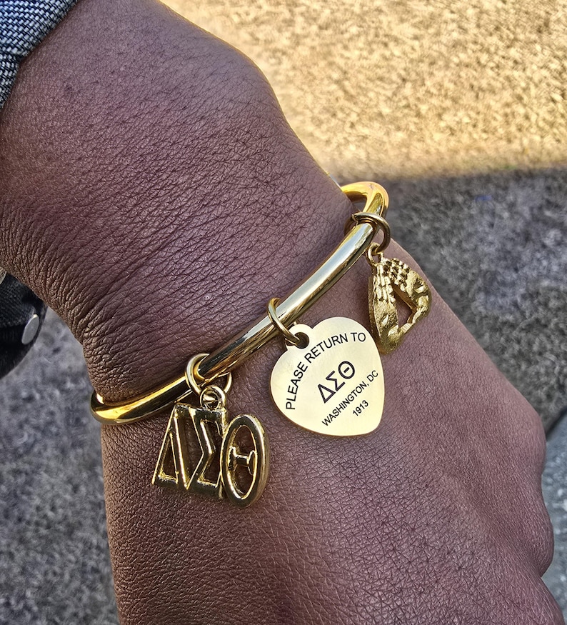 May include: Gold charm bracelet with a heart-shaped charm that says "Please return to Delta Sigma Theta, Washington, DC 1913". The bracelet also has a charm with the letters "DST" and a charm with a handprint.