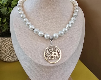 Op de afbeelding: Een parelketting met een gouden hanger. De ketting heeft grote, ronde, witte parels en zilveren accenten. De gouden hanger heeft de woorden "DELTA SIGMA THETA" in een cirkelvormig ontwerp. De ketting wordt op een mannequin getoond.