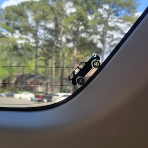 May include: A black car decal is affixed to a car window. The decal is a silhouette of a car, angled as if driving uphill. The background shows trees, a blue sky, and other vehicles.
