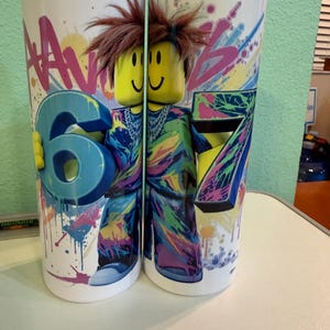 May include: Two white tumblers with colorful graffiti-style designs. One tumbler features a large blue number 6, the other a number 7. Both have a Lego-like figure with brown hair and a smiling face. The tumblers are decorated with paint splatters.
