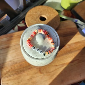 Puede incluir: Un recipiente de cerámica blanca con un brazalete de cuentas de colores que deletrea "BOO". El brazalete está hecho de cuentas rojas, naranjas, blancas y negras.