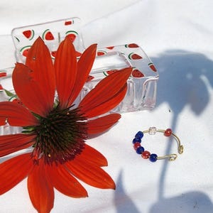 May include: A vibrant image featuring a red flower, a clear hair clip with watermelon slices, and a gold-toned ring adorned with red and blue beads. The items are arranged on a white surface, with the flower's shadow visible.