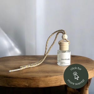 May include: A clear glass car air freshener bottle with a light wood cap and a beige braided hanging cord. The white label features the text boardwalk and homebody lush. A dark green circular graphic with the text click for more scents is present.