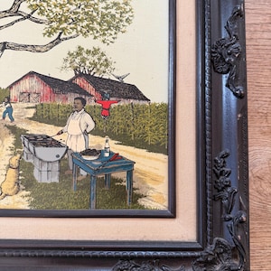 Peut inclure: Une peinture encadr&eacute;e repr&eacute;sentant une sc&egrave;ne rurale avec un homme faisant griller des aliments sur un barbecue au charbon de bois. Il y a deux granges rouges en arri&egrave;re-plan, un &eacute;pouvantail et un arbre. La peinture est dans un cadre brun fonc&eacute; avec des d&eacute;tails orn&eacute;s.