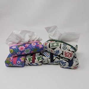 May include: A set of six tissue box covers. Two covers feature a floral pattern in shades of pink, purple, and green. The other four covers feature a mahjong tile pattern in shades of green, red, and blue. The covers are made of fabric and have a quilted texture.