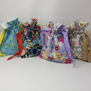 May include: Four drawstring bags in various floral patterns. The bags feature blue, red, purple, and beige floral prints. Each bag has a contrasting ribbon drawstring in yellow, red, purple, and beige. The bags are made of fabric and are gathered at the top.