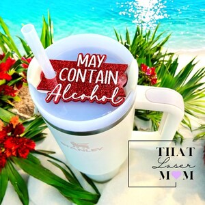 May include: White travel mug with a red glitter lid that says "May Contain Alcohol". The mug is on a white sandy beach with green plants and blue water in the background.