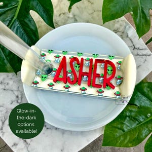 Op de afbeelding: Een op maat gemaakte, rechthoekige tray met de naam "ASHER" in felrode letters. De tray heeft een herhalend patroon van groene alien gezichten en vliegende schotels. De tekst "Glow-in-the-dark options available!" is ook zichtbaar.