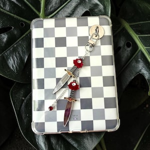 Op de afbeelding: Een zilverkleurige metalen sleutelhanger met twee dolkbedels, elk versierd met een rode roos en een kleine witte schedel. De dolken zijn verbonden door een zilveren ketting met een enkele rode kristallen kraal aan het uiteinde, aan een ring.