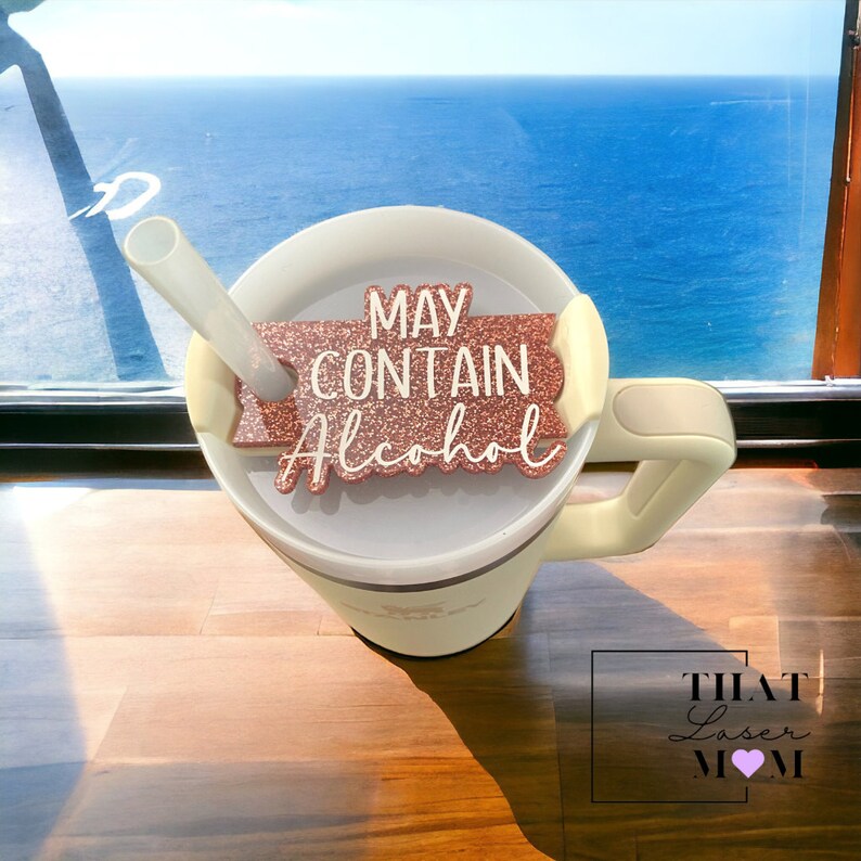 May include: A white travel mug with a pink glitter  "May Contain Alcohol"  sticker attached to the side. The mug has a straw and a handle. The mug is sitting on a wooden table with a view of the ocean in the background.