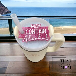 May include: White travel mug with a handle and a pink glitter sticker that says "May Contain Alcohol". The mug is on a wooden surface with a blue ocean and sky in the background.