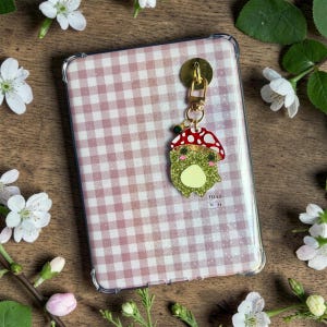 Pode incluir: Uma capa de telefone transparente com um padrão xadrez rosa e branco. Um pingente dourado com um topo em forma de cogumelo e um design de sapo verde brilhante está preso à capa. Flores brancas e vegetação circundam a capa.