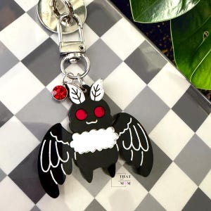 May include: A black and white moth keychain featuring red eyes and a small red rhinestone charm attached to a silver-toned metal lobster clasp. The moth has a white fluffy chest area and white wing details. The text That Laser Mom is visible.