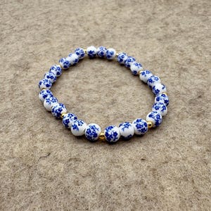 Puede incluir: Un brazalete de cuentas de porcelana blancas y azules con un diseño floral. El brazalete presenta cuentas redondas con un patrón floral azul y espaciadores dorados. El brazalete se muestra sobre una superficie de tono neutro.