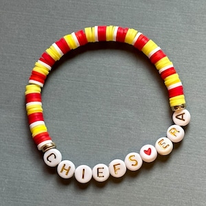 Könnte beinhalten: Ein weißes Perlenarmband mit den Worten "CHIEFS" und "ERA" in goldenen Buchstaben. Das Armband hat außerdem eine rote Herzperle und einen rot-weiß-gelb gestreiften Abschnitt.