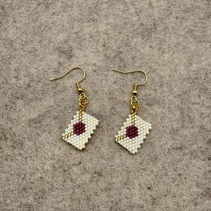May include: A pair of handmade earrings featuring a white and burgundy beaded design. The earrings have a rectangular shape with a burgundy circle in the center and gold-colored ear wires. The earrings are displayed on a neutral-colored surface.