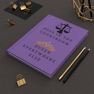 May include: Purple notebook with the text "Boss in the courtroom, Queen everywhere else". The notebook has a gold crown and a black scale of justice graphic.