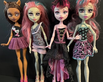 May include: Four fashion dolls standing in a row. The first has brown hair and a black and purple outfit. The second has pink and blue hair and a patterned dress. The third has black and pink hair and a black dress. The fourth has pink and blue hair and a black and pink patterned dress.