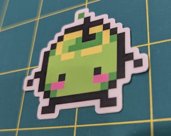 May include: A pixelated sticker of a green creature with pink cheeks and black outlines. The creature has a yellow and green head and a white border. The sticker is on a green cutting mat.