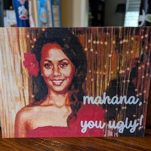 May include: A rectangular print featuring a portrait of a person with dark hair and a red flower behind their ear, wearing a red garment. The background shows golden vertical light strands. The text reads: mahana, you ugly!