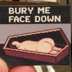 May include: A rectangular sticker with the text "BURY ME FACE DOWN" in white pixelated font. The image below depicts a pixelated person lying face down in a coffin. The coffin is dark red with a black border, and the background is light purple.