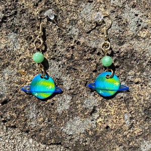 Puede incluir: Un par de pendientes hechos a mano con un diseño de planeta. Cada pendiente tiene un gancho dorado, una pequeña cuenta verde y un colgante de planeta con tonos azules, verdes y amarillos, y anillos azules. Los pendientes se exhiben sobre una superficie texturizada de tonos neutros.