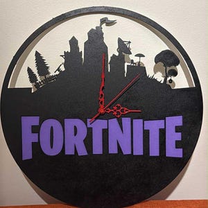 May include: Black and purple Fortnite-themed wall clock. The clock features a silhouette of a cityscape and the word "FORTNITE" in purple. Red clock hands complete the design. The clock is round and appears to be made of wood.