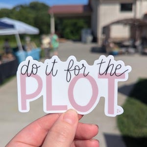 May include: A white sticker with the phrase "do it for the PLOT" in a mix of cursive and block letters. The word "PLOT" is in a light pink color. The sticker is held in a hand against a blurred outdoor background.
