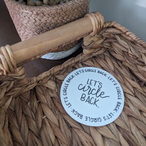 May include: A white round sticker with the text "LET'S circle BACK" in black lettering. The sticker is placed on a woven basket.