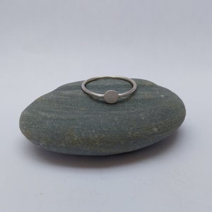 May include: A simple silver ring with a round, flat top. The ring is resting on a grey rock.