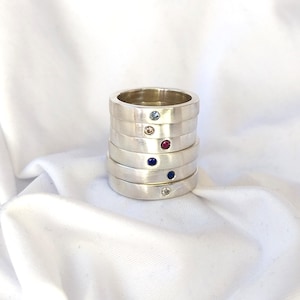 May include: Stack of five silver rings with a different coloured gemstone set in each ring. The gemstones are blue, red, pink, and clear.