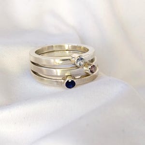 May include: Three silver rings stacked together, each featuring a different coloured gemstone. The top ring has a light blue stone, the middle ring a mauve stone, and the bottom ring a dark blue stone. The rings rest on white fabric.