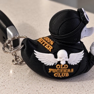 May include: A black and white 3D-printed duck keychain wearing a motorcycle helmet and goggles. The duck features a white skull with wings, the text 100% BITTER, and the text OLD FUCKER CLUB in orange. The duck is gesturing with a middle finger.