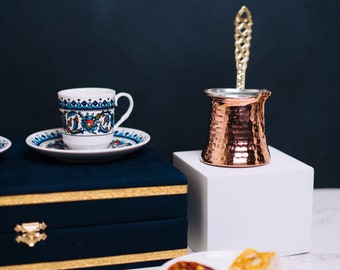 May include: A traditional Turkish coffee set featuring a small, patterned cup and saucer, a copper coffee pot, and a decorative spoon. The set is displayed with a plate of pastries and a dark blue box.