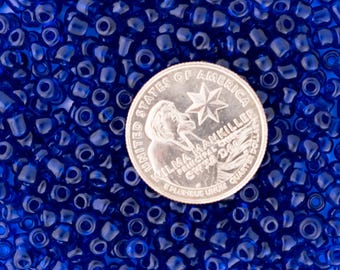 Puede incluir: Una imagen de primer plano de un montón de pequeñas cuentas de semillas azules translúcidas. Se incluye una moneda de 25 centavos de dólar estadounidense para la escala.
