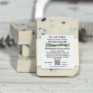 May include: Three bars of white soap with black flecks. The top bar has a label that reads "the velvet tallow Rustic & Hand Crafted Beef Tallow & Goat Milk Rosemary Eucalyptus Elements: Saponified Grass-Fed Beef Tallow, Coconut Oil Castor Oil, Kaolin Clay, Activated Charcoal, Lard, Essential Oil Blend of (Rosemary, Eucalyptus, Lavender, Cedarwood) Net Weight oz Made in Slidell, LA www.TheVelvetTallow.com"