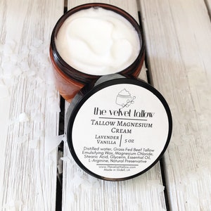 May include: An open jar of The Velvet Tallow Tallow Magnesium Cream, with a white, creamy texture. The jar is brown with a black lid, which displays the product name, ingredients, and size: 5 oz. The cream is scented with lavender and vanilla.