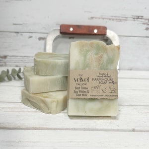 May include: A stack of four green and white farmhouse soap bars with a brown paper label that reads "the velvet TALLOW Rustic & Hand-Milled FARMHOUSE SOAP Wt 5oz Beef Tallow Egg Whites & Goat Milk French Green Clay & Turmeric".