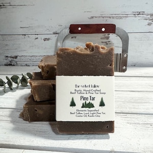 May include: A bar of brown soap with a white label that reads "the velvet tallow Rustic, Hand-Crafted Beef Tallow & Pine Tar Soap Pine Tar Elements: Saponified Beef Tallow, Lard, Light Pine Tar, Castor Oil, Kaolin Clay"