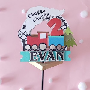 Op de afbeelding: Een rode, blauwe en groene trein caketopper met een witte wolk waarop "Chugga Chugga" staat en een houten basis. De trein heeft een nummer 2 aan de zijkant en de naam "EVAN" aan de voorkant.