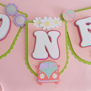Peut inclure: Une bannière rose et verte avec le mot "ONE" en lettres blanches. La bannière est décorée de marguerites, de signes de paix et d'un van vintage.