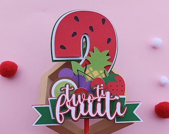 May include: A two-tier cake topper featuring a watermelon shaped number two with a variety of fruit decorations. The topper is decorated with a green banner that says "two-ti frutti".