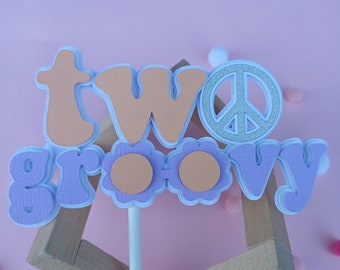 May include: Purple and orange cake topper with the words "two groovy" and a peace sign.