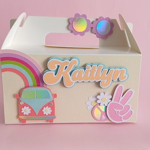 Puede incluir: Una caja de regalo blanca decorada con un arcoíris, un signo de paz y una furgoneta rosa. La caja tiene el nombre "Kaitlyn" y está decorada con flores y gafas de sol iridiscentes.