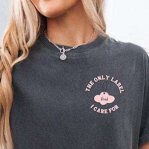 May include: Dark gray t-shirt with the text "THE ONLY LABEL I CARE FOR" in a pink circle with a cloud graphic. The model is wearing denim shorts and a cream-colored cardigan. A decorative pumpkin and woven basket are in the background.