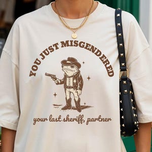 Puede incluir: Camiseta de color marfil con un gráfico marrón de un sheriff rana sosteniendo una pistola, con el texto "YOU JUST MISGENDERED" arriba y "your last sheriff, partner" abajo. La camiseta la lleva una persona.